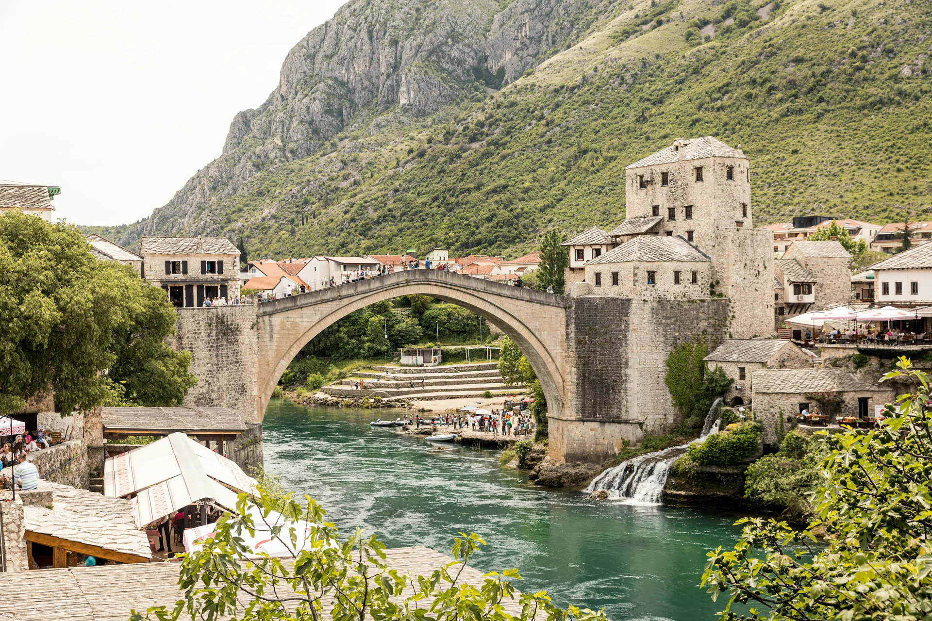 Stari Most, Mostar's rebuilt Ottoman arch bridge, spanning the Neretva river with the Old Town tower and a small waterfall beside it